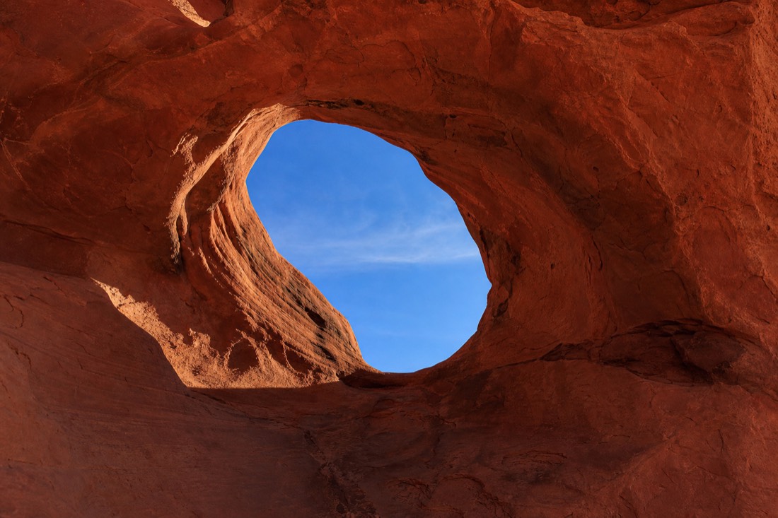Sun's Eye, a natural sandstone opening framing a patch of blue sky through curved red rock in the backcountry