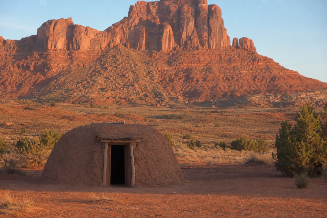 A traditional Navajo hogan on the valley floor with red buttes rising behind it in soft morning light