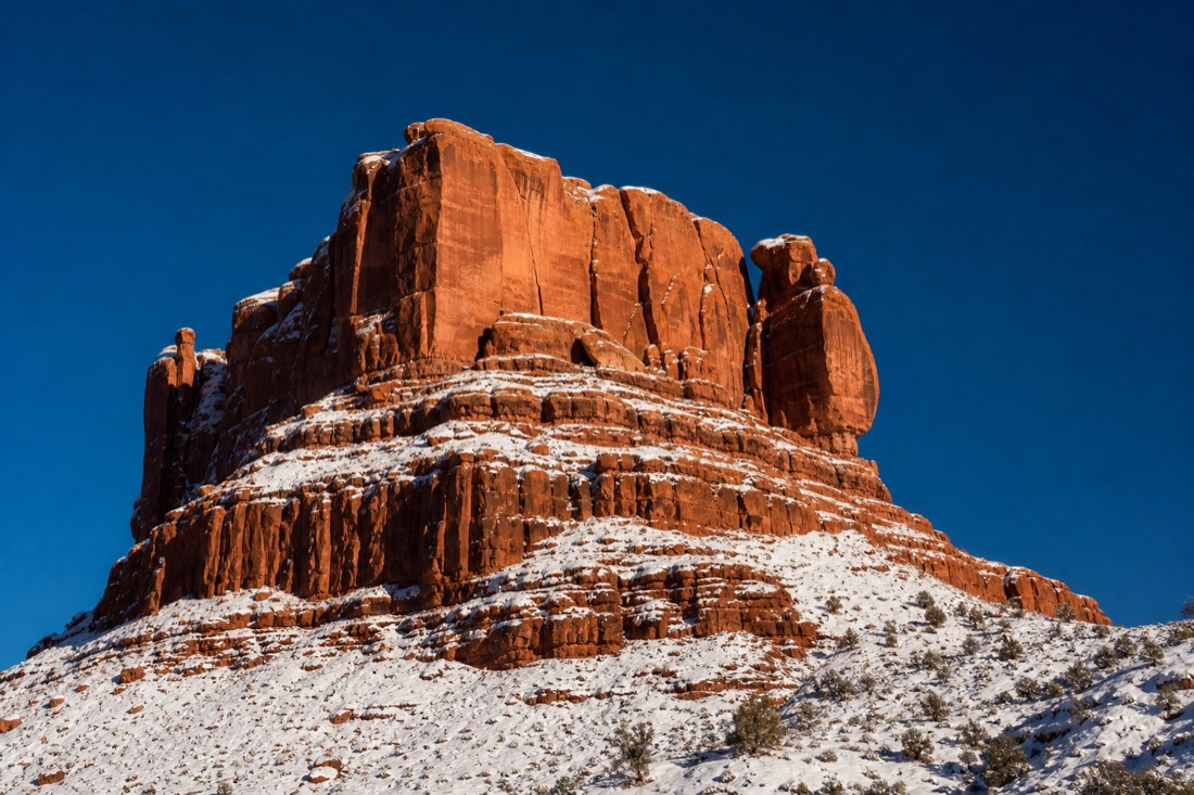 Fresh snow clinging to the ledges of a towering red sandstone butte against a crisp deep blue sky