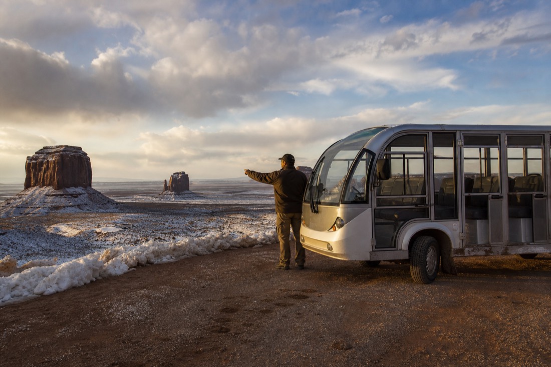 An enclosed tour vehicle parked at a snow-touched overlook with a guide pointing toward distant winter buttes
