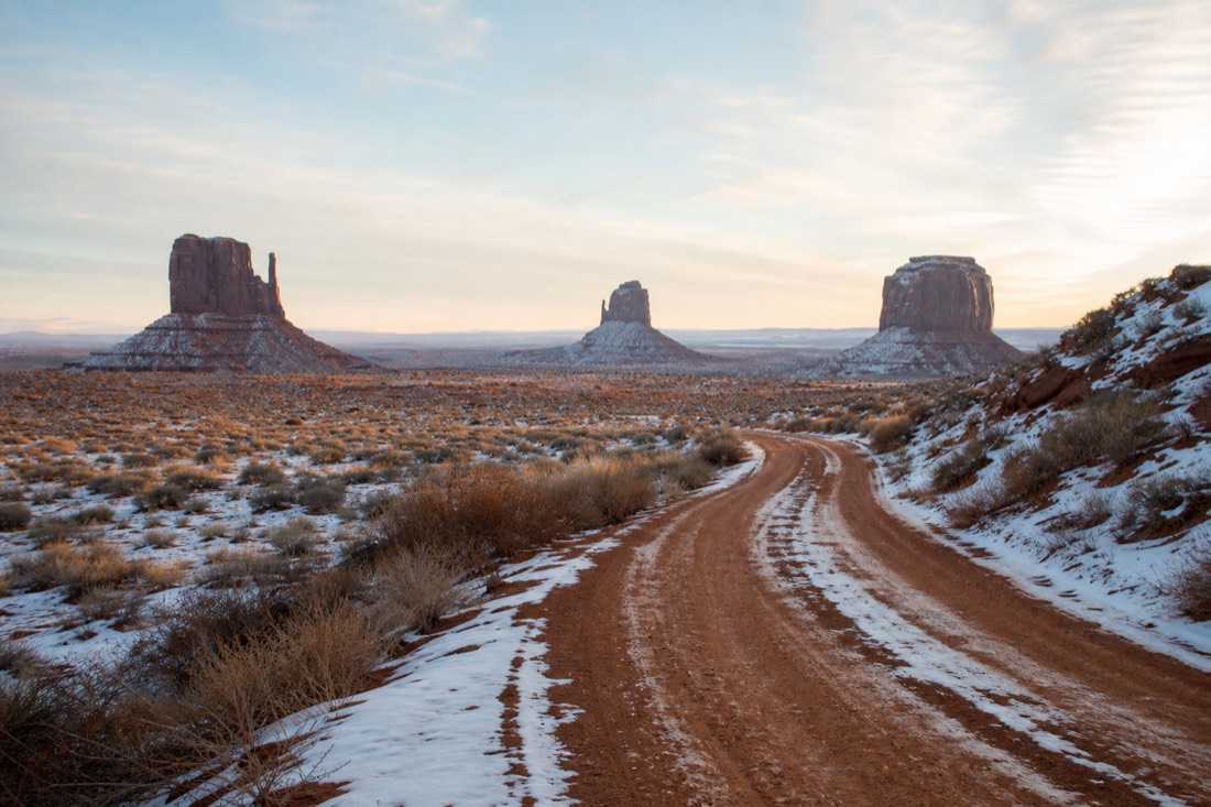 An empty snow-dusted red dirt road curving toward the iconic buttes under soft winter light with no other visitors