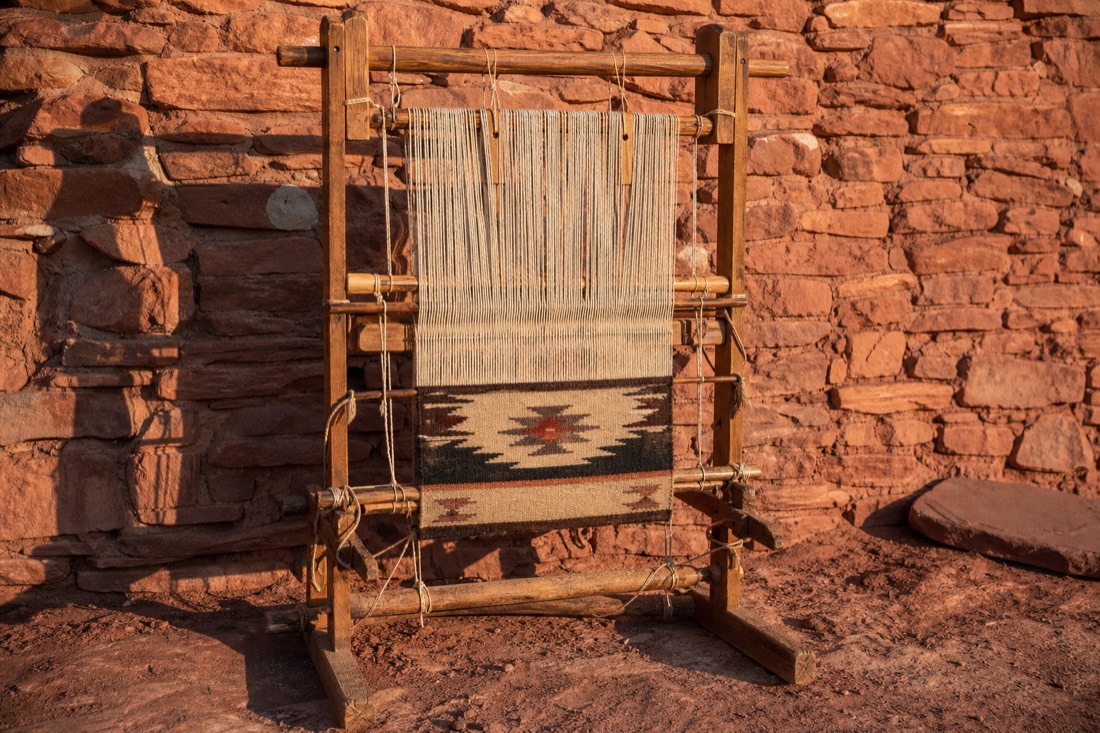 An upright wooden Navajo loom with a half-finished wool rug set against a red sandstone wall