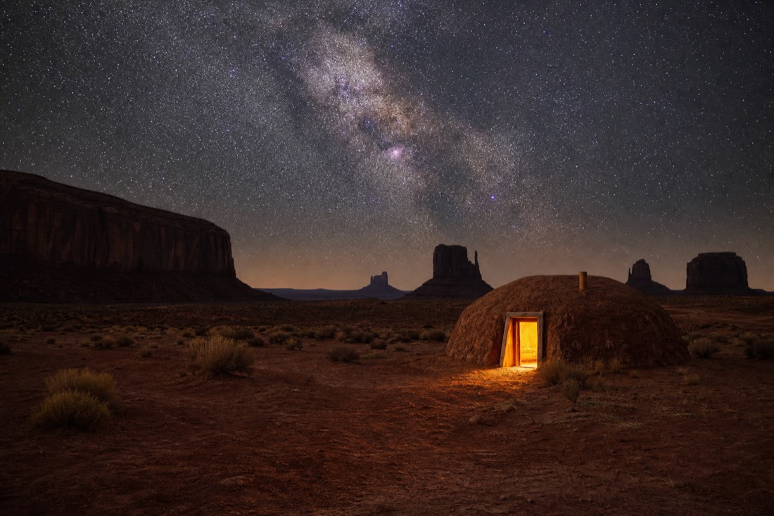 A traditional hogan glowing warm from its doorway on the valley floor with the Milky Way overhead and buttes silhouetted around it