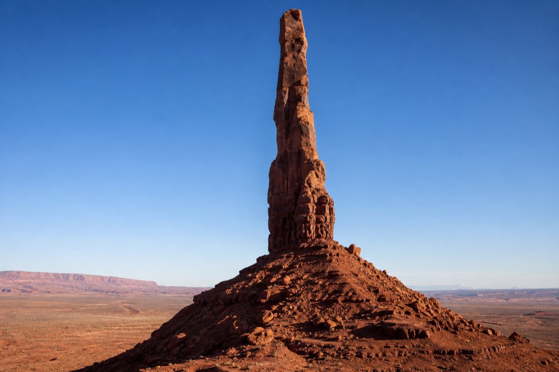 A single tall thin sandstone spire rising alone from the red desert floor against a clear blue Monument Valley sky
