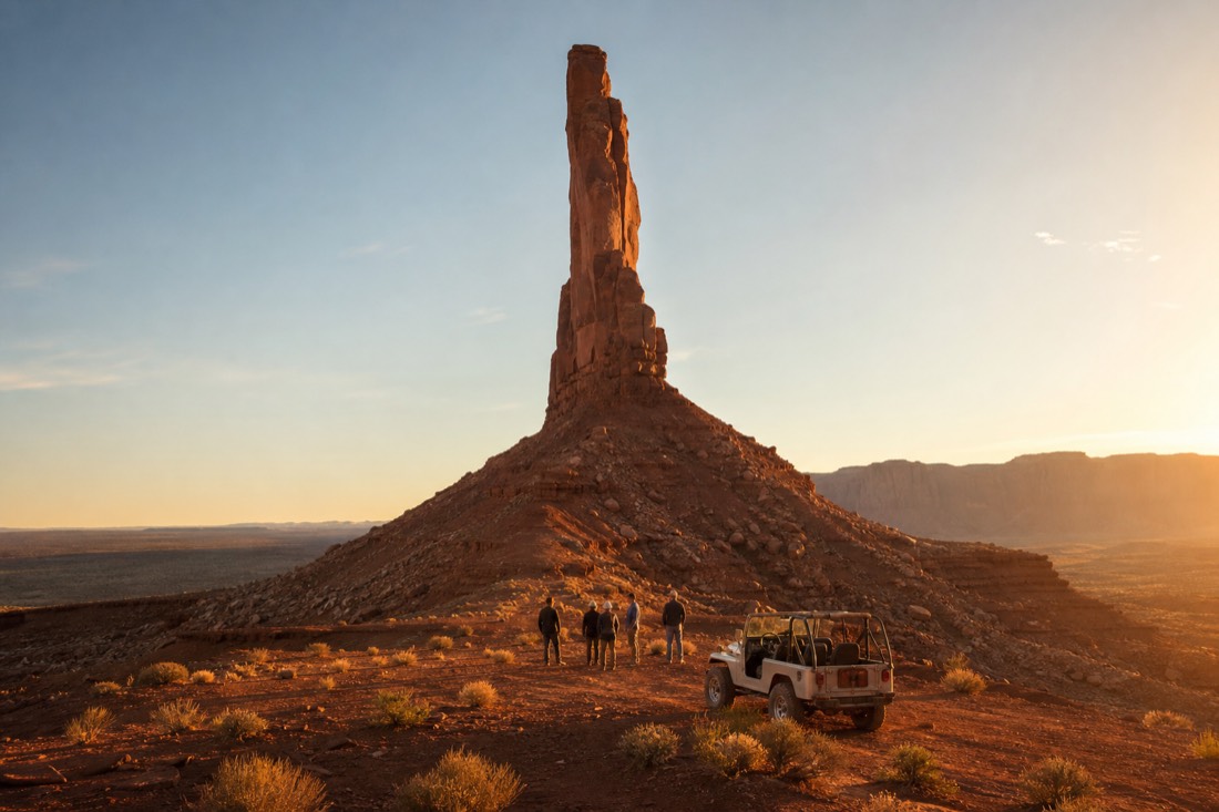 Golden hour light on a lone slender rock spire with an open-air jeep and small group at its base in the Monument Valley backcountry
