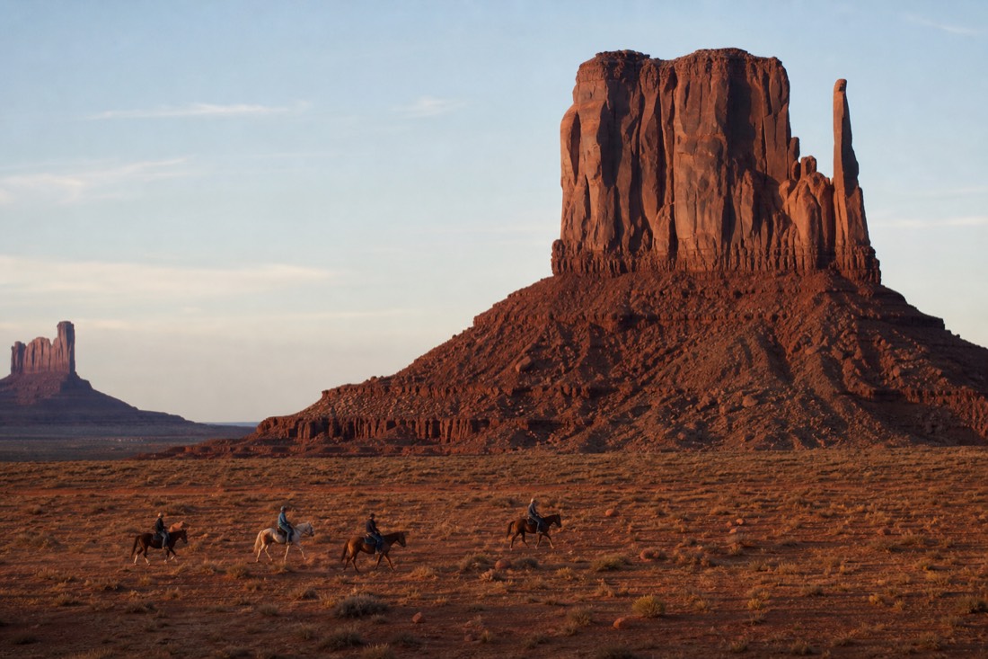 Horseback riders crossing the open red desert floor beneath the towering buttes of Monument Valley