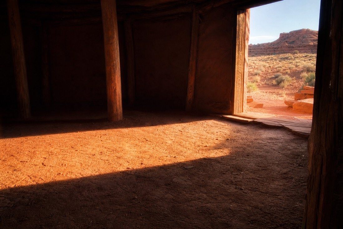 Low morning sunlight streaming through the open east-facing doorway of a hogan onto a packed-earth floor