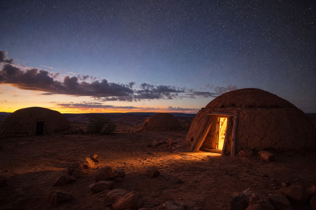 A cluster of hogans at dusk with warm lantern light glowing from a doorway under a star-filled desert sky