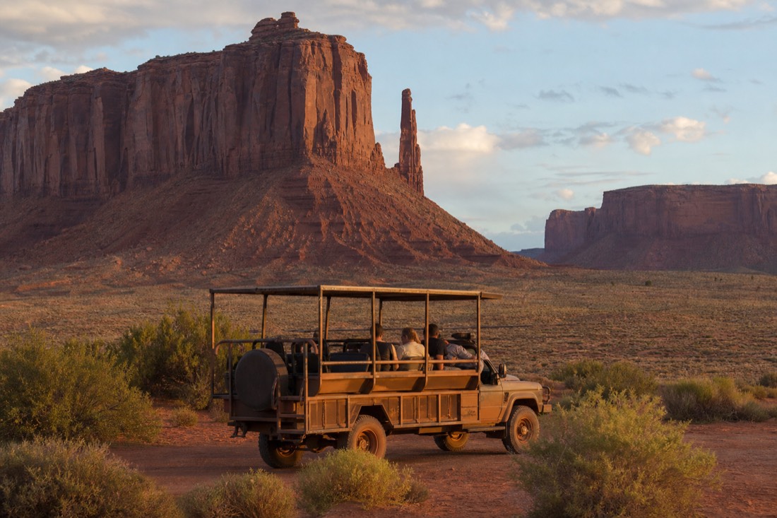 A small group in an open-air Navajo tour vehicle pausing beneath towering red buttes on the valley floor in warm light