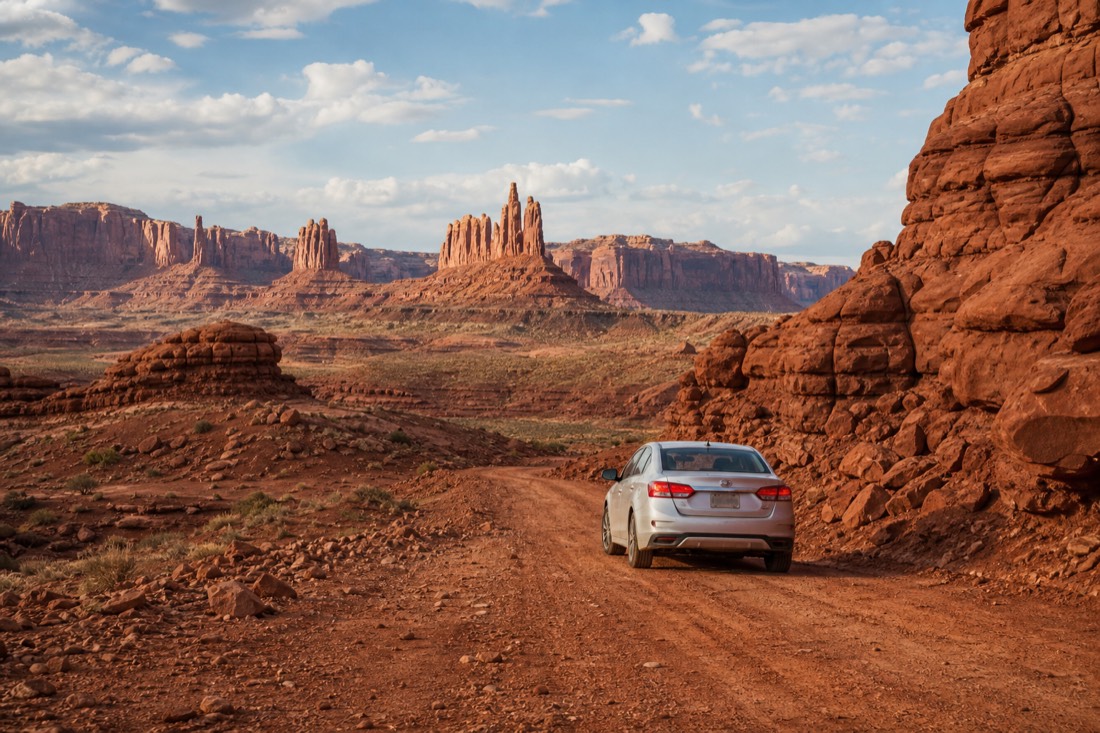 A lone rental car on the rough dirt Valley Drive with red spires rising in the distance under a bright sky
