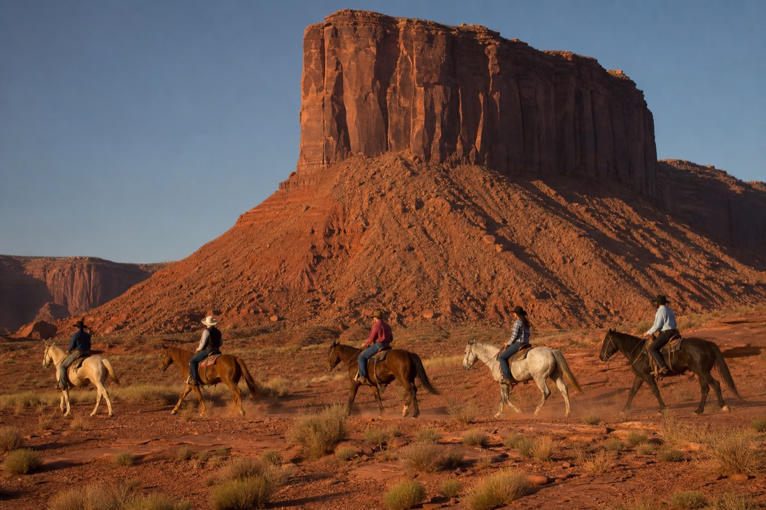 Riders on horseback crossing the open red valley floor beneath a massive butte in late afternoon light