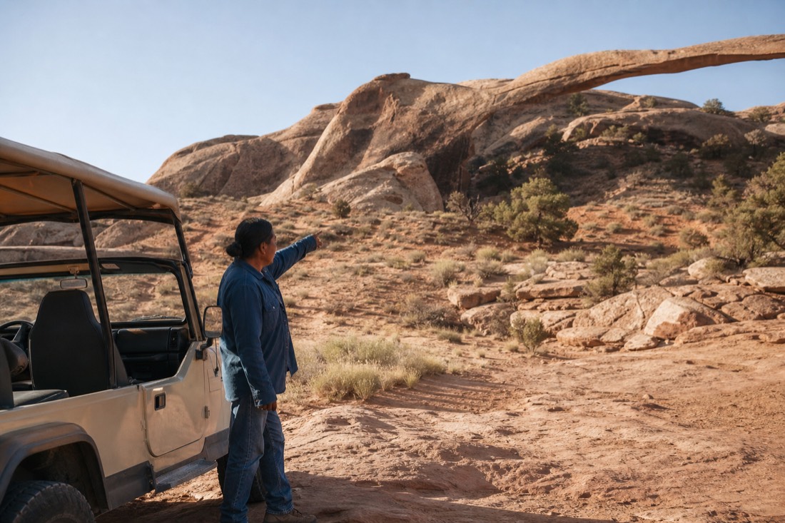 A Navajo guide standing beside an open-air jeep gesturing toward a sandstone arch in the quiet backcountry