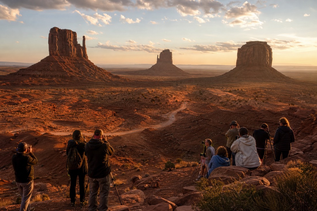 A wide golden-hour view of the Mitten Buttes and valley floor with a small tour group taking photos from an overlook