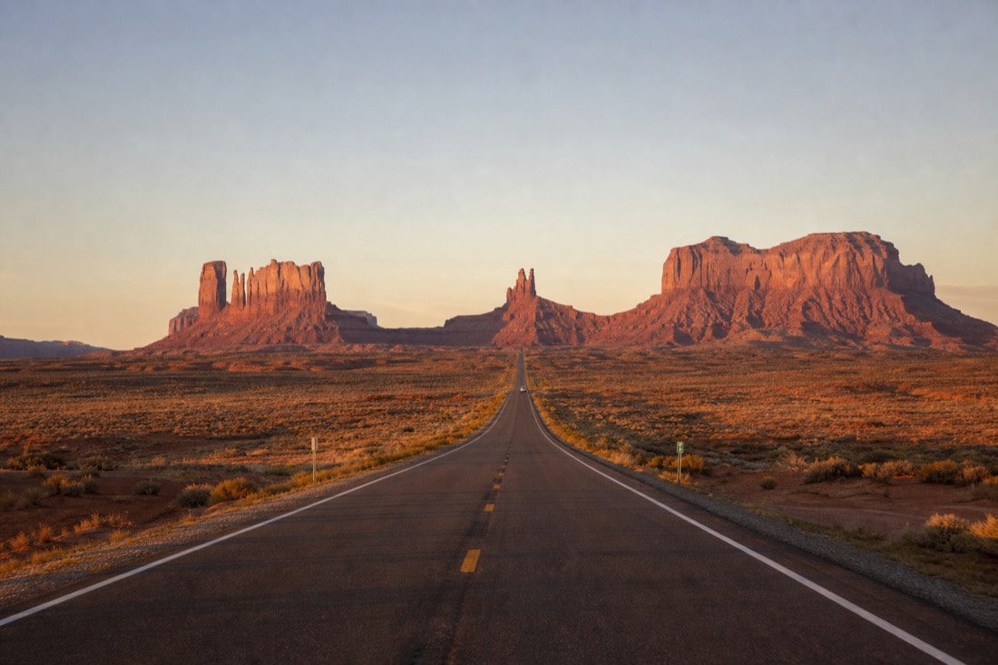 US-163 highway running straight toward Monument Valley's buttes at golden hour
