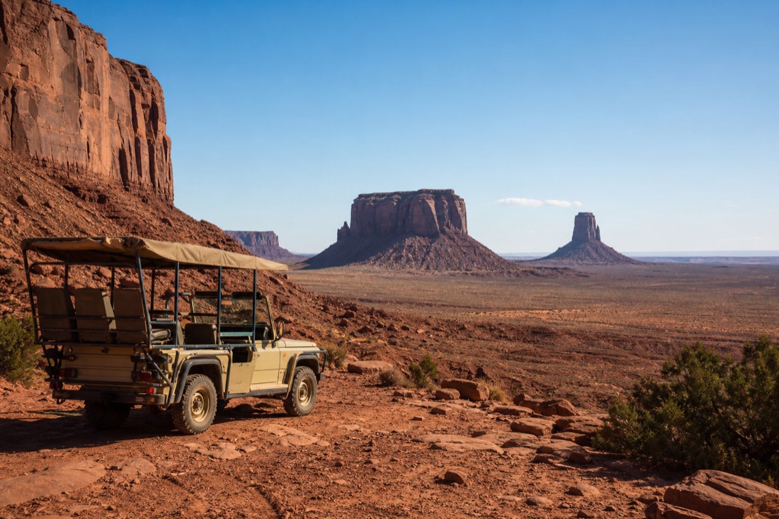 A Navajo-guided open jeep parked at a viewpoint over the valley floor at mid-morning, buttes sharp under a clear high sun