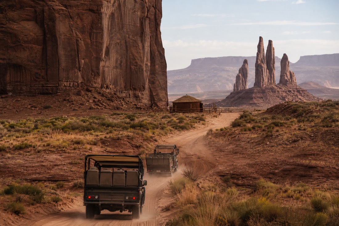Two open-air tour vehicles on a sandy backcountry track winding toward a hogan and distant spires in Mystery Valley