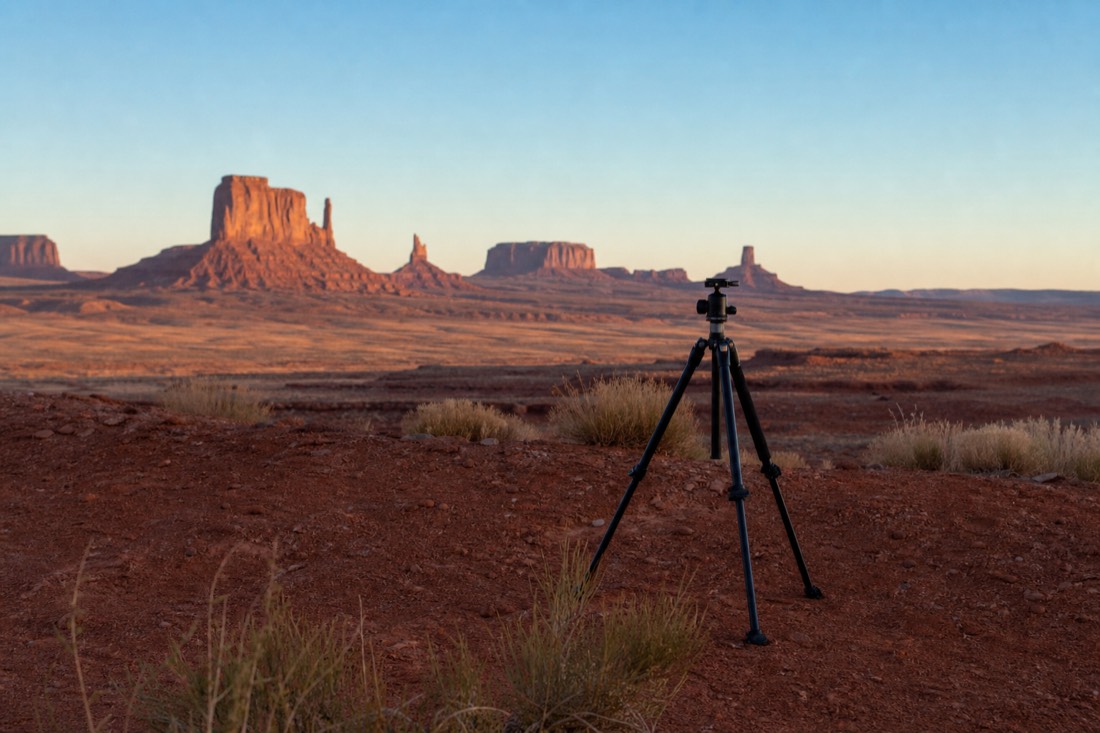 An empty camera tripod set on red dirt facing the Mittens as first light warms the eastern rock faces