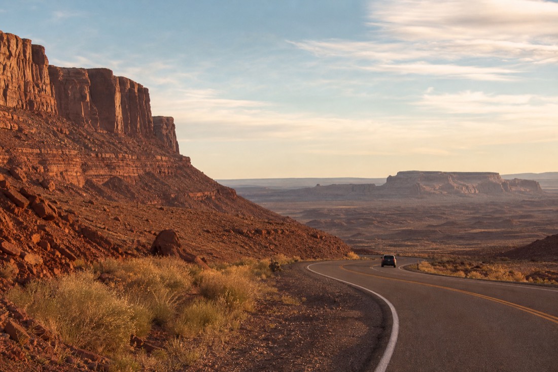 A lone car on a distant desert highway winding through red-rock mesa country under a wide sky