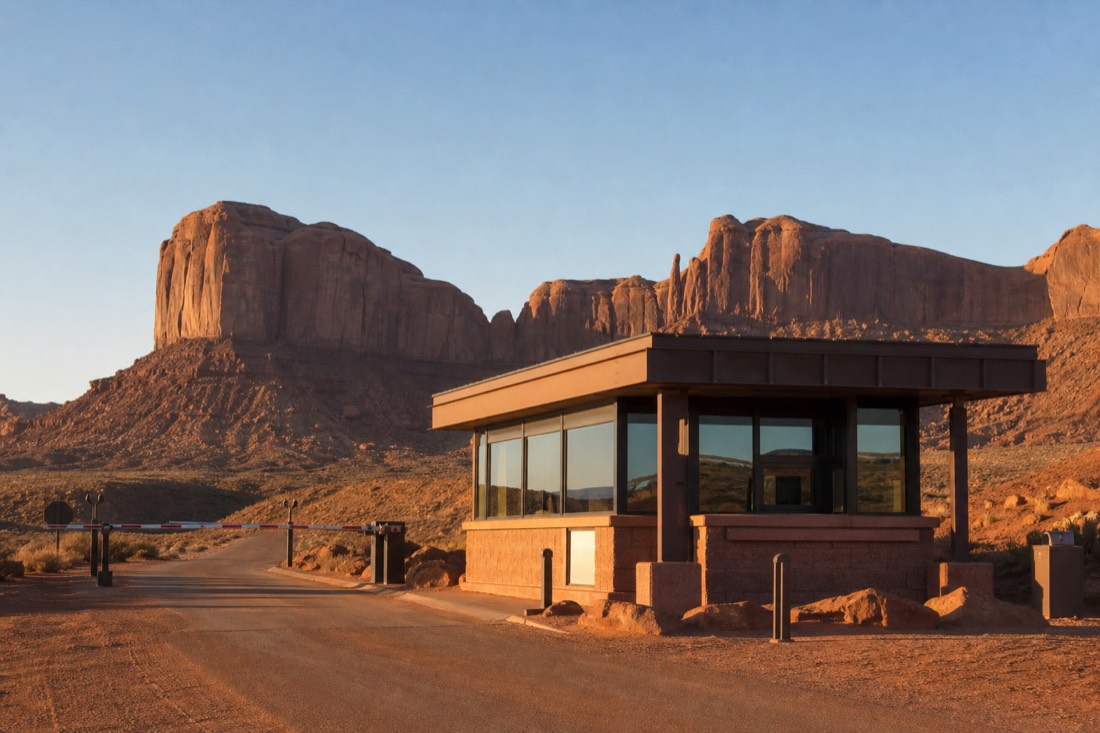 A simple park entrance station on open red desert with towering buttes behind it, no branded signage or logos