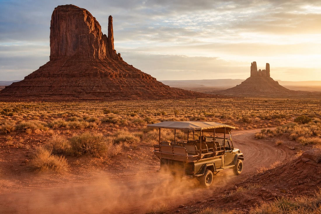An open-air guided jeep on the red dirt loop road kicking up dust with Merrick Butte and the Mittens rising behind at golden hour