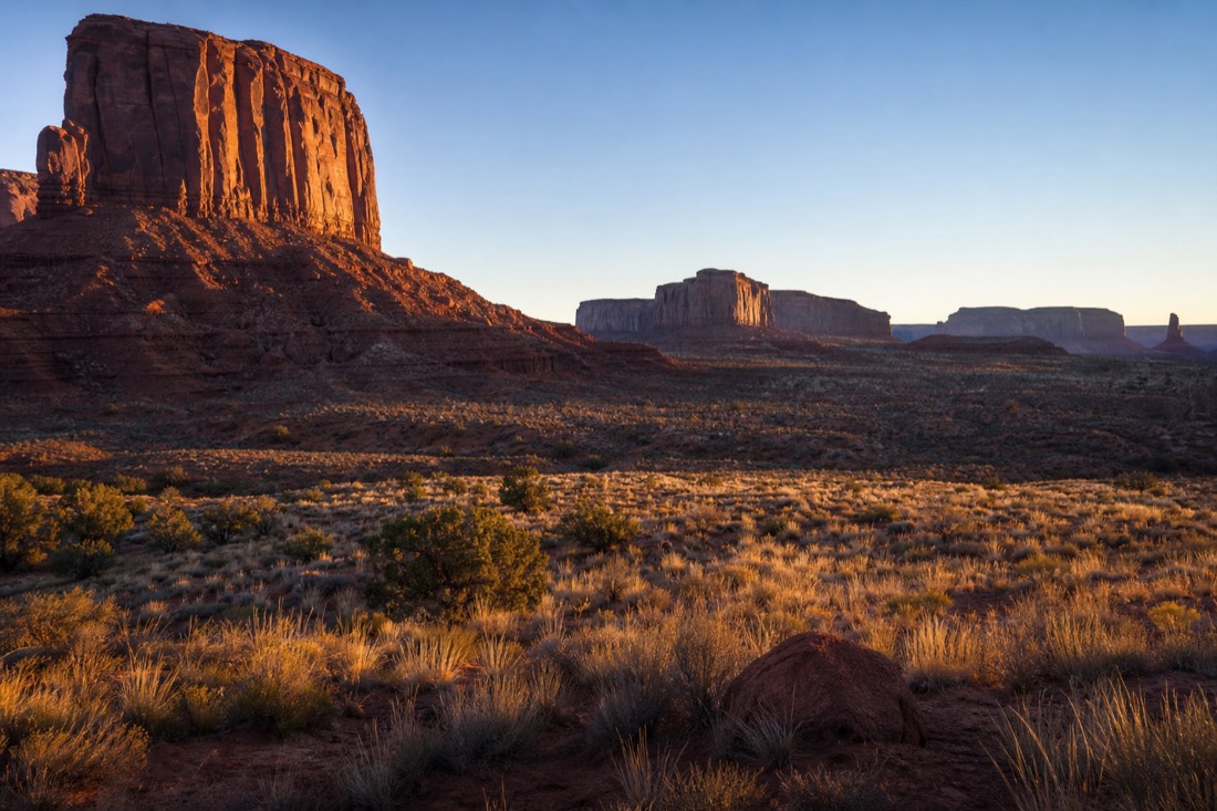 Low autumn sun raking across the buttes, long blue shadows stretching over golden desert grass and red sand