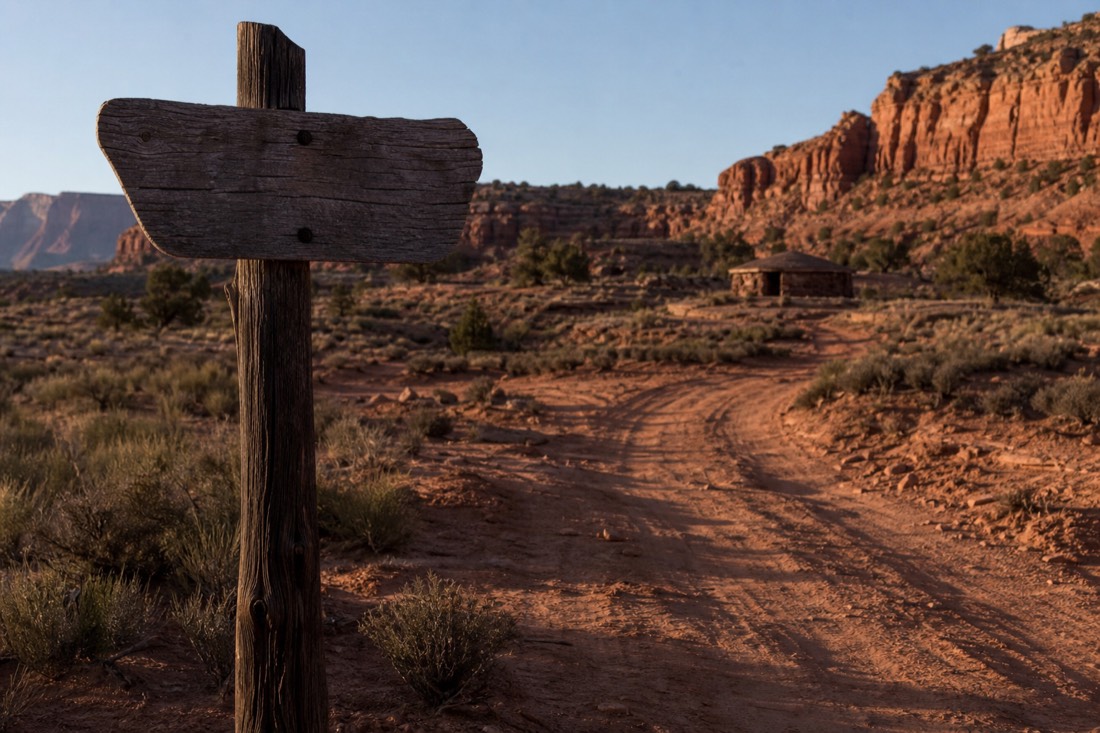 A weathered wooden sign post and marked dirt track winding into red-rock backcountry, a traditional hogan roofline in the distance