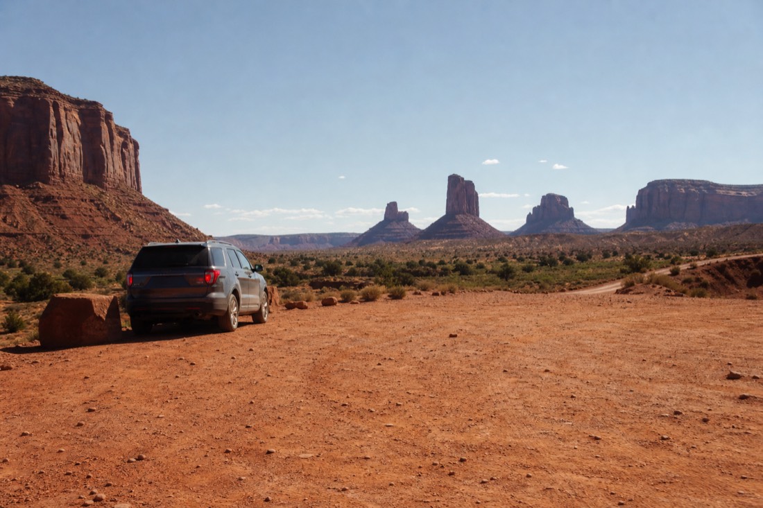 A red-dirt turnout on the Valley Drive at mid-morning with a parked SUV and the Mittens rising in clear cool light, no people