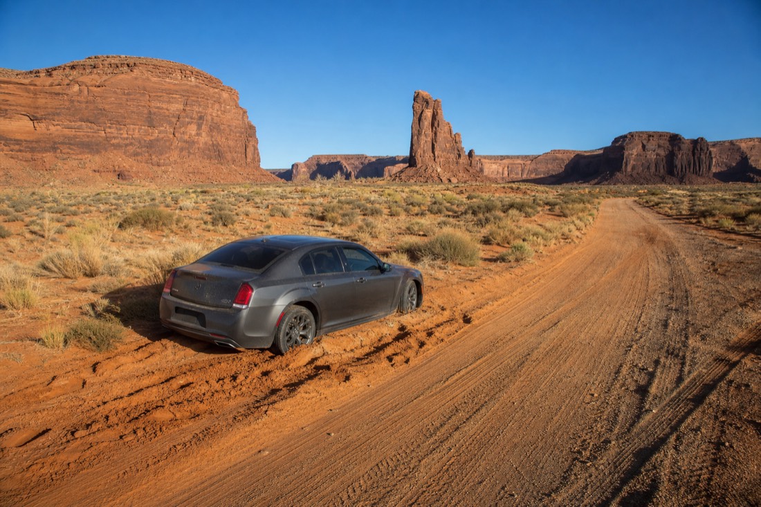 A low sedan stopped in deep sand on the washboard loop road, tire tracks veering off the marked track, buttes behind under bright sky