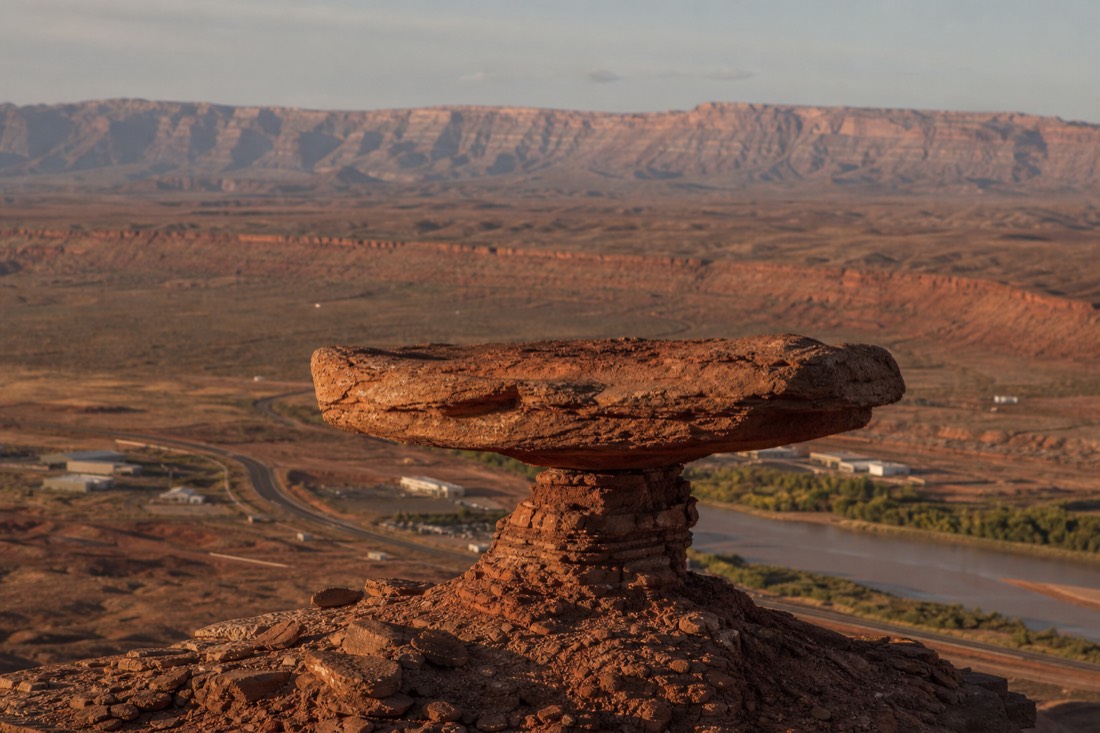 A flat sombrero-shaped rock disc balanced on a slim stone pedestal above a small highway settlement beside a brown desert river