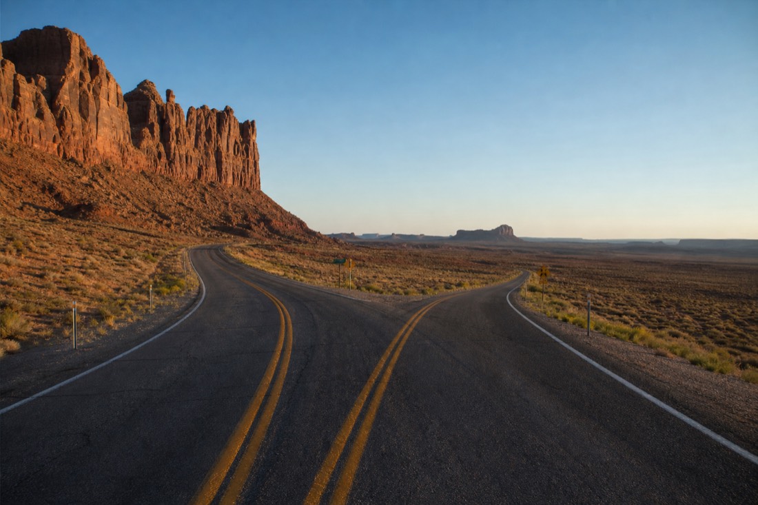 A straight desert highway splitting two directions, red Utah cliffs to one side and open Arizona flats with distant buttes to the other