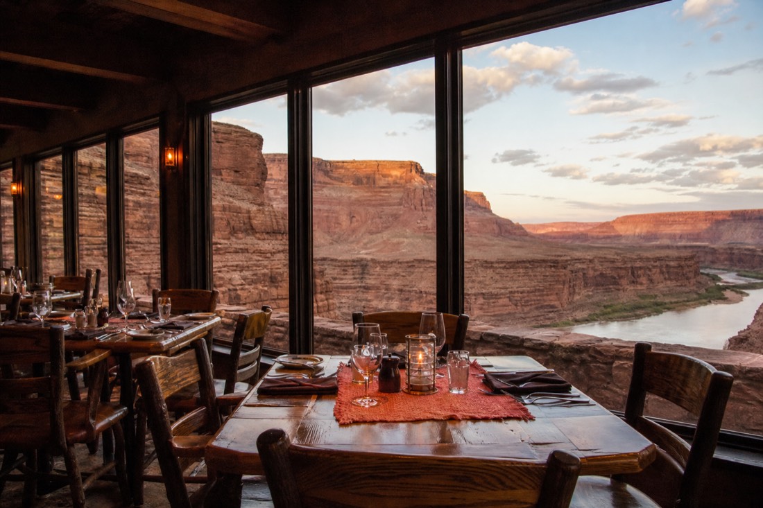 A rustic desert inn dining room with big windows framing red canyon walls and the river below, empty tables set for evening