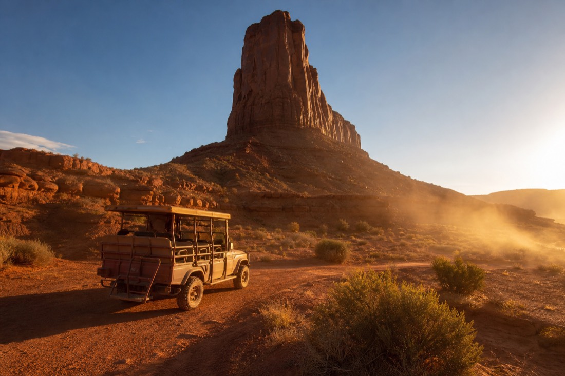 An open-air tour vehicle on red backcountry sand beneath a towering sandstone spire, dust and long shadows in warm light