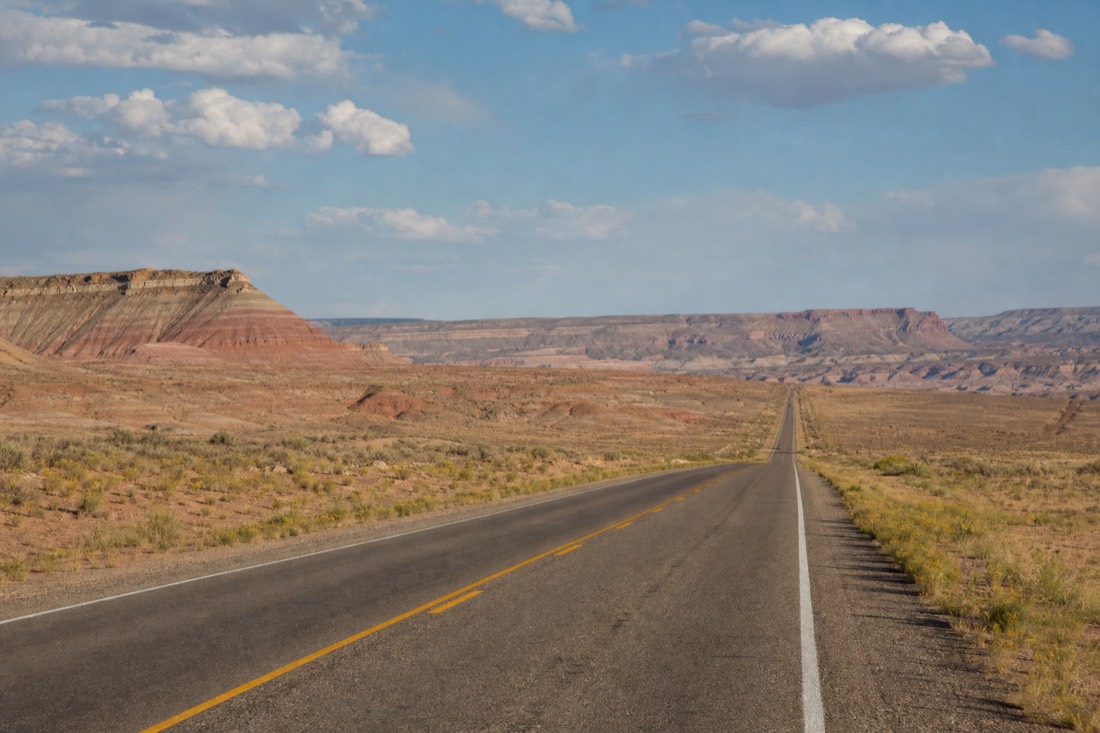 A long straight desert highway crossing bands of pink and gray painted-desert badlands under a big sky