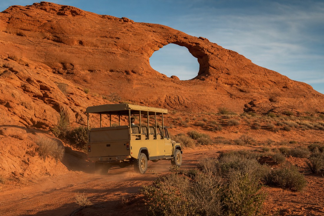 An open-air Navajo tour jeep passing the Ear of the Wind arch in the red-dune backcountry beyond the loop road