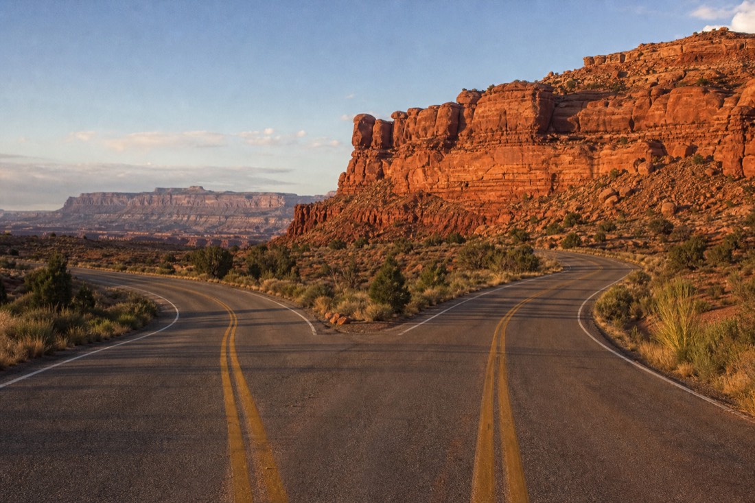 A road sign junction where a desert highway splits southwest, red cliffs rising on both sides in late afternoon light
