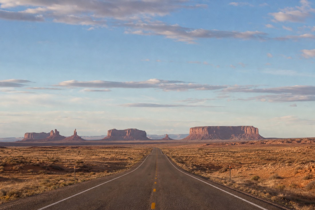 An empty paved highway cutting straight across open red-and-tan Navajo high desert toward distant flat-topped mesas under a wide sky