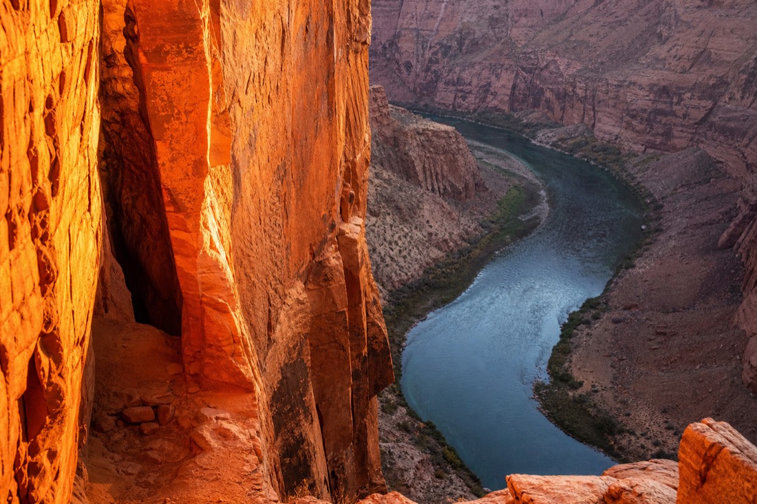 A narrow glowing orange slot canyon wall beside a sweeping blue-green river bend carved deep into pale sandstone