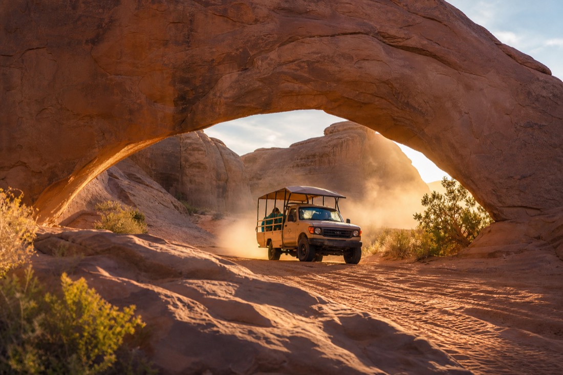 An open-air Navajo tour vehicle passing beneath a natural sandstone arch on red backcountry dunes, dust hanging in golden light