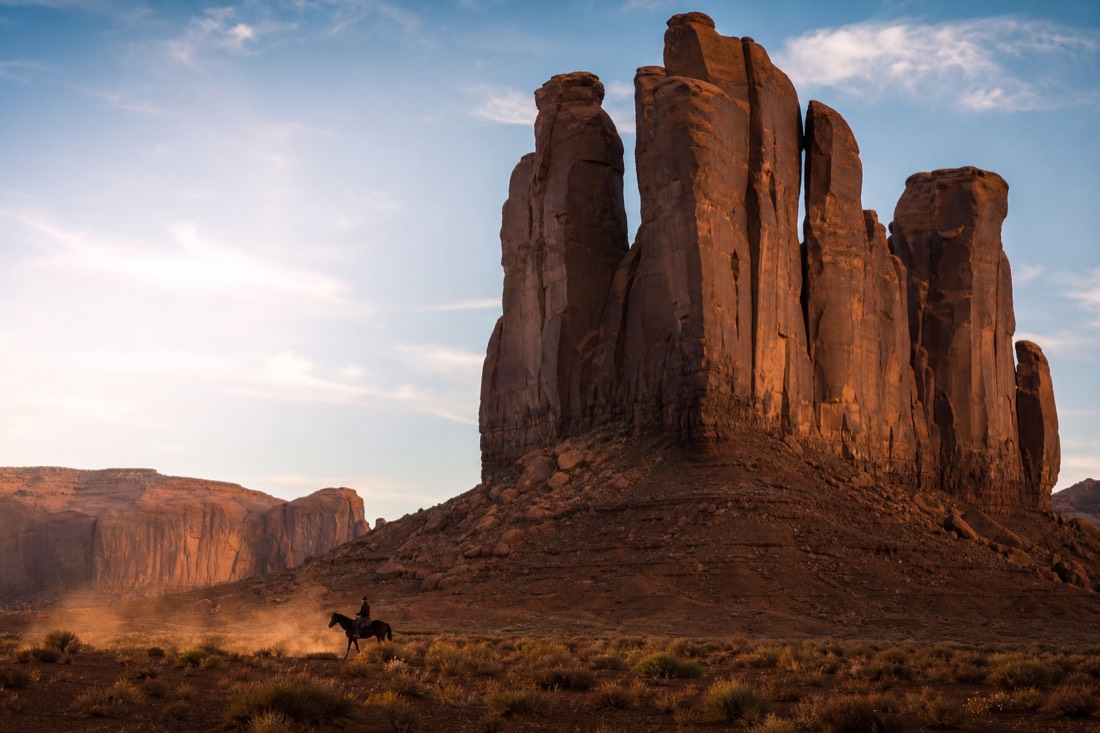 A lone rider silhouetted on horseback beneath enormous sandstone spires in dramatic low sunlight
