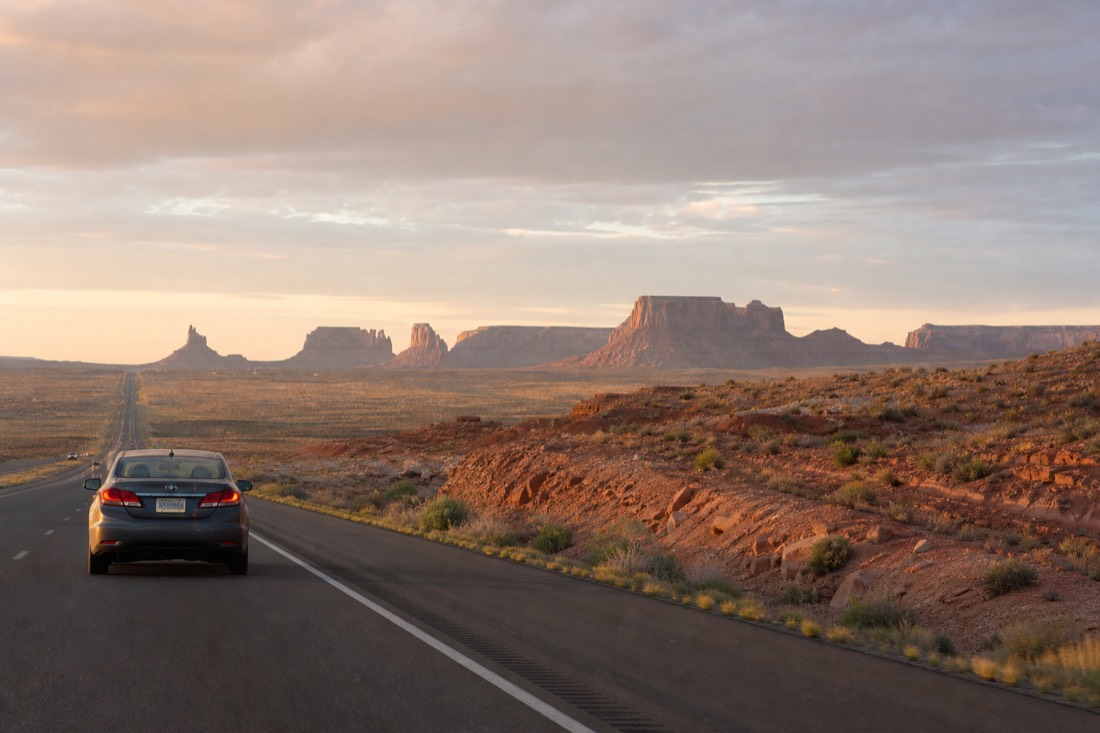A rental car on an open interstate leaving a city behind toward distant desert buttes at dawn