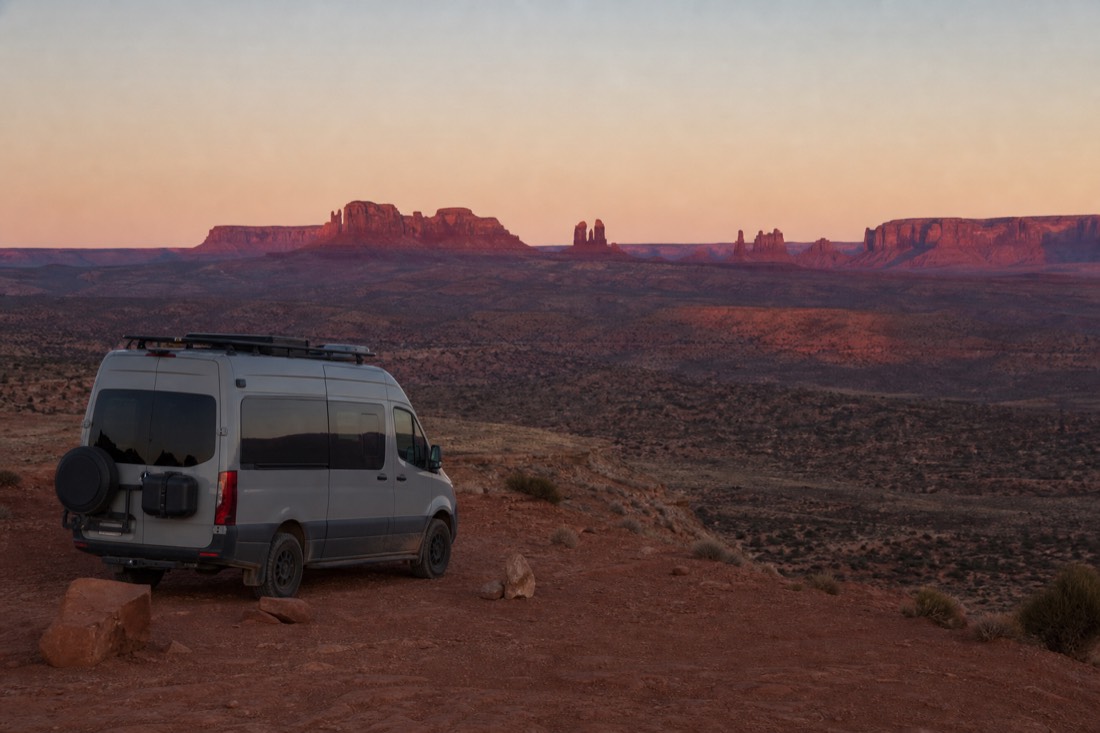 A campervan parked at a desert overlook at first light with red spires glowing on the far horizon
