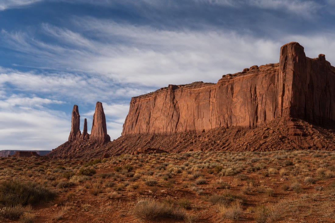 Wide isolated sandstone buttes and slender spires rising from open scrubland under a big Utah sky, no roads or people in view