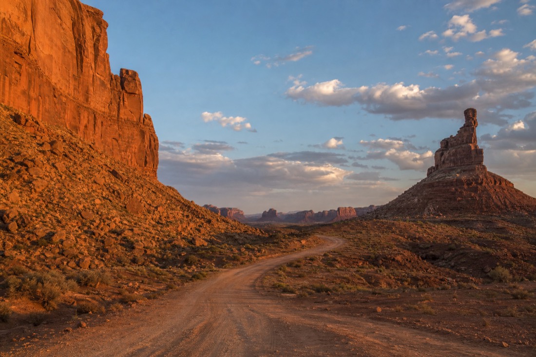 A lone empty dirt road curving between tall red rock formations in Valley of the Gods, no vehicles or signage