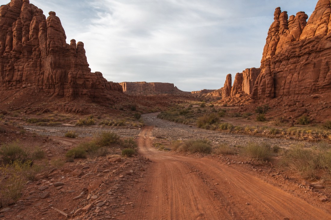 A dirt back road crossing a shallow dry desert wash between sandstone spires, tire tracks pressed into red clay