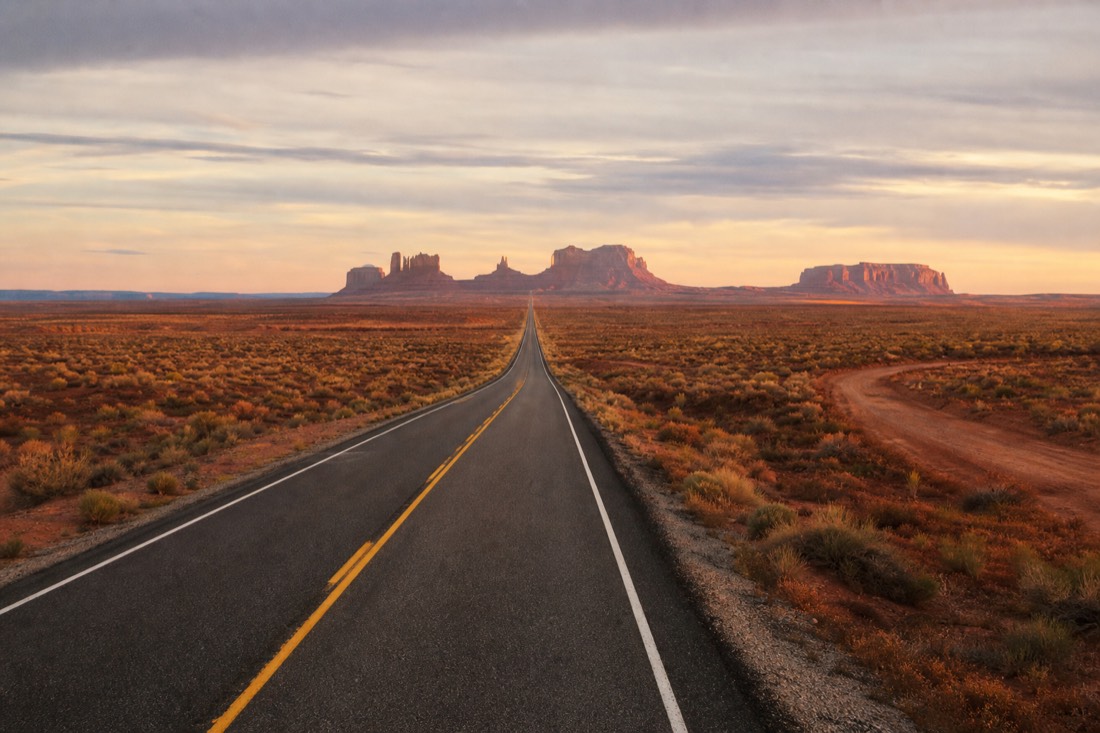 US-163 running dead straight toward distant Monument Valley buttes at dusk, a side dirt turnoff branching into open desert