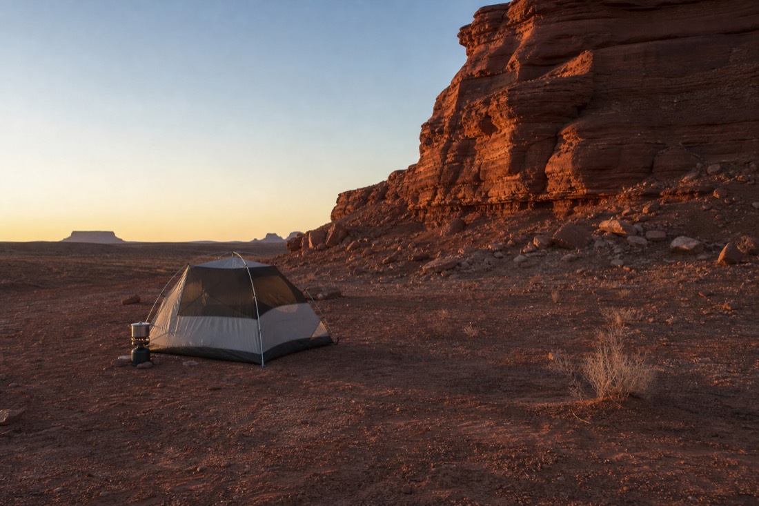 A small tent pitched on bare dirt beside a sandstone butte at dusk, a camp stove and no facilities in sight, empty desert all around