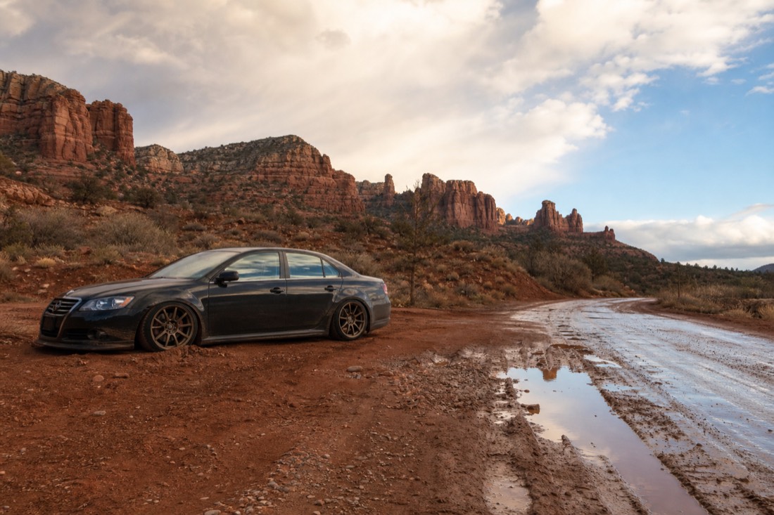 A long low-clearance sedan stopped at the muddy edge of a rain-soaked dirt road below red buttes, ruts filled with water