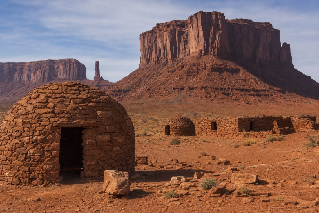 Weathered stone hogans and low sandstone buildings below the towering red buttes of Monument Valley