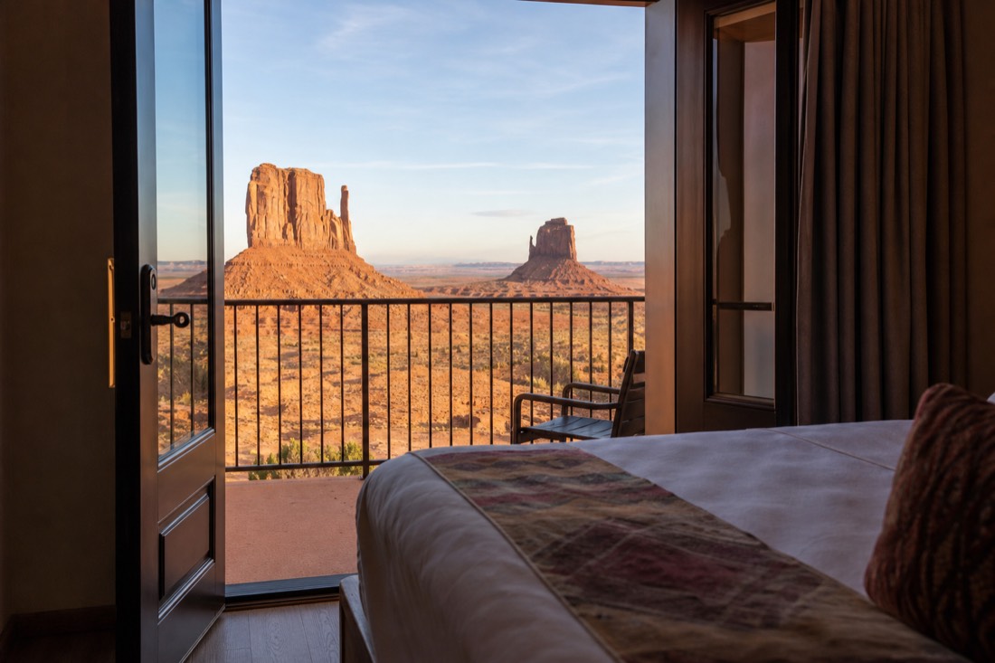 A made hotel bed beside an open balcony door framing the sunlit Mitten buttes in early morning light