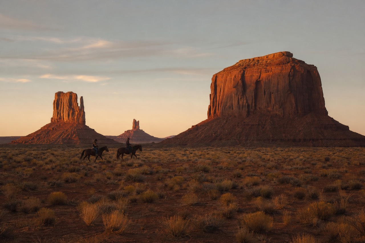 Riders on horseback crossing the red desert floor of Monument Valley below the buttes at golden hour